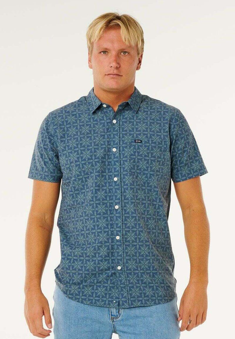 Jonge man met kort blond haar, gekleed in een blauw shirt met korte mouwen en patroon, en lichtblauwe spijkerbroek, staand tegen een effen achtergrond.