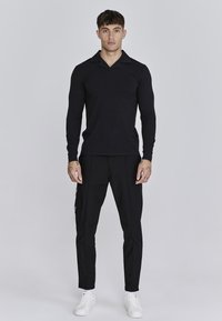 Camisa polo negra de manga larga con cuello puntiagudo, combinada con pantalones negros ajustados y zapatillas blancas. Las características incluyen un ajuste slim y un diseño minimalista.