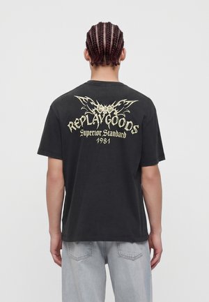 Oseba s kitami, gledana od zadaj, oblečena v črno majico s svetlo rumenim besedilom in dizajnom, na katerem piše "REPLAY GOODS Superior Standard 1981".