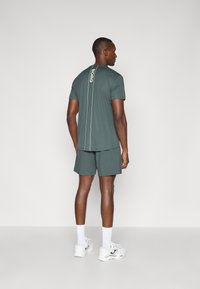 Σκούρο πράσινο αθλητικό μπλουζάκι με λευκή κάθετη ρίγα και λογότυπο στο πίσω μέρος, σε συνδυασμό με ταιριαστά σορτς και λευκά αθλητικά παπούτσια.