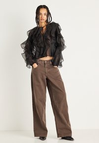 Blusa nera trasparente con strati di volant e un fiocco davanti, abbinata a pantaloni in denim marroni larghi. L'outfit presenta un design rilassato e una vestibilità dritta.