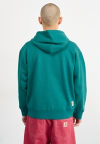 Sweatshirt à capuche teal avec une coupe décontractée, des poignets et un ourlet côtelés. Logo de la marque en bas à gauche, associé à un pantalon rose. Texture de coton lisse.
