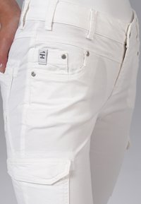 Pantalon en denim blanc avec une coupe slim, des poches devant et des poches cargo, ainsi qu'une petite étiquette de marque sur la taille avec des rivets en argent.