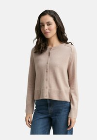 Frau mit welligem, dunklem Haar, die einen beigefarbenen, knopfartigen Cardigan und eine blaue Jeans trägt, im Vordergrund einer schlichten, hellen Hintergrunds.