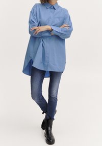 Camicia blu a maniche lunghe con colletto, tasca frontale e orlo curvo abbinata a jeans scuri e stivali neri alla caviglia.