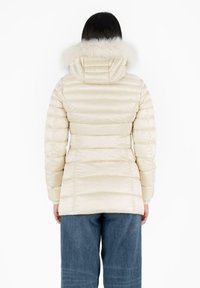 Chaqueta tipo puffer en un beige pálido con acolchado horizontal, que presenta una capucha de piel sintética. Textura suave y silueta ajustada, que cae por debajo de las caderas.