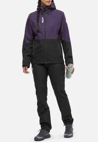 Lila und schwarze wasserdichte Jacke mit einem Reißverschluss vorne, Kapuze und Seitentaschen, kombiniert mit schwarzen Hosen und Wanderschuhen, die eine grüne Wasserflasche hält.