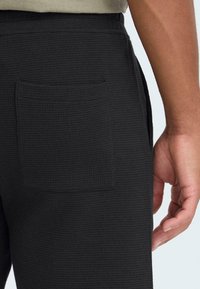 Pantalon texturé noir avec une poche arrière, une ceinture lisse et une main visible reposant sur le côté.