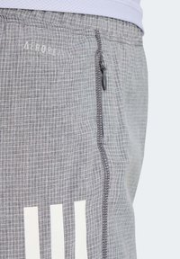 Pantalones cortos deportivos grises con un patrón de cuadros, bolsillo con cremallera lateral y rayas blancas de la marca en la parte inferior. Hechos de un material transpirable.