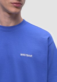 Niebieski sweter z fakturowanej tkaniny ma okrągły dekolt i małe białe haftowane logo "WRSTBHVR" na piersi.