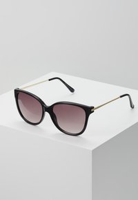 Schwarze Cat-Eye-Sonnenbrille mit einem Verlaufsglas, ausgestattet mit goldenen Metallelementen an den Bügeln und einem glänzenden schwarzen Rahmen.