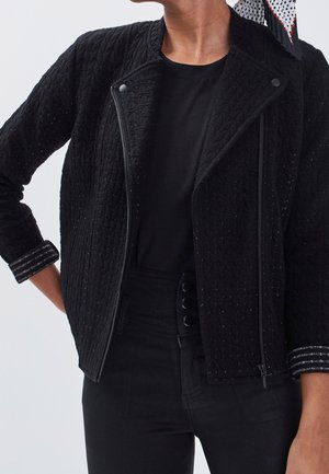 Vrouw draagt een zwarte textuurritsjas over een zwart topje en zwarte broek met hoge taille, met een stippen sjaal in het haar geknoopt.