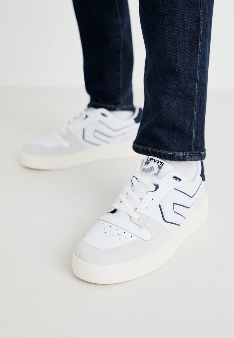 Vita och grå sneakers med övre i slätt läder, marinblå detaljer och en texturerad tunga med perforeringar för andningsförmåga.