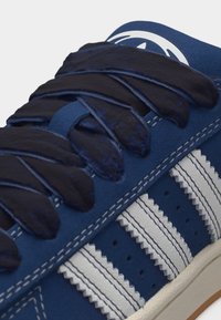 Μπλε αθλητικό παπούτσι με σκούρα μπλε κορδόνια, πλευρικές λωρίδες με άσπρες ζιγκ-ζαγκ ρίγες και ραμμένη άκρη στη σόλα.