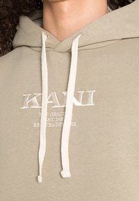 Khaki mikina s kapucí s bílými šňůrkami, vyšívané logo a text "KAN" na hrudi, hladká textura a ležérní design.