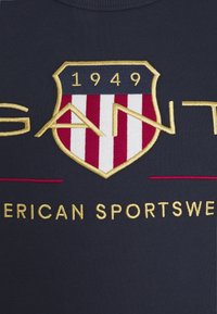 Felpa blu navy con scollo rotondo caratterizzata da un logo a scudo ricamato con strisce rosse e bianche, lettering dorato e dettaglio "1949".