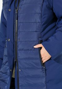 Giacca isolata blu navy con una combinazione di pannelli trapuntati e tessuto liscio, dotata di tasche con zip e chiusure a bottoni a pressione.