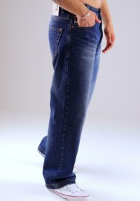 Mann in lockeren blauen Jeans und weißen Segeltuchschuhen, der mit einer Hand in der Tasche vor einem schlichten Hintergrund steht.