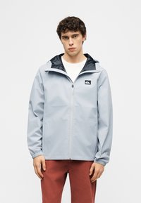 Νεαρός με σγουρά μαλλιά φοράει ένα ανοιχτό γκρι μπουφάν με φερμουάρ και κουκούλα και κόκκινο παντελόνι, στέκεται μπροστά σε μια απλή φόντο.