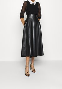 Svart midi kjol i konstläder med hög midja, tillsammans med en svart topp med korta genomskinliga ärmar och en vit krage.