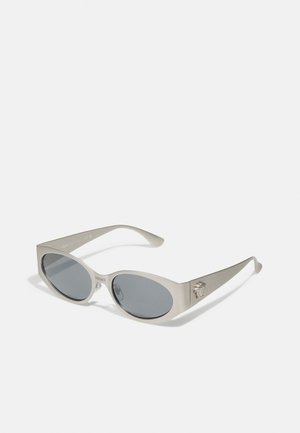 Occhiali da sole ovali oversize con montatura in metallo argento, lenti grigie scure e logo di leone in rilievo sul lato; design elegante e minimale.