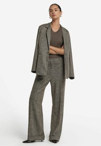 Donna che indossa un completo in scozzese marrone con pantaloni a gamba larga e un blazer abbinato drappeggiato sulle spalle, abbinato a un top a maniche corte a coste.