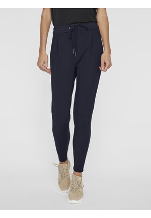 Pantalon jogger bleu marine slim avec taille à cordon porté avec des baskets beiges, montré de la moitié du torse aux pieds sur fond clair.