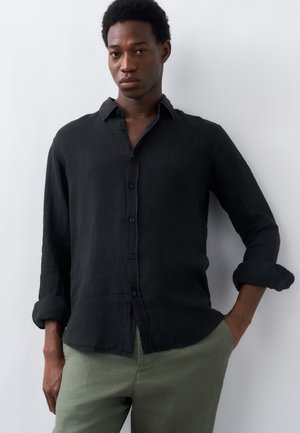 Młody mężczyzna w czarnej koszuli na guziki z podwiniętymi rękawami i zielonych spodniach, stojący na tle jednolitego białego tła.