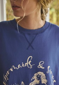 Nahaufnahme einer Person, die einen blauen Pullover mit dem Text "Mermaids & Sirens" und einer Meerjungfrau-Grafik trägt, mit sichtbar lockeren blonden Haarsträhnen.