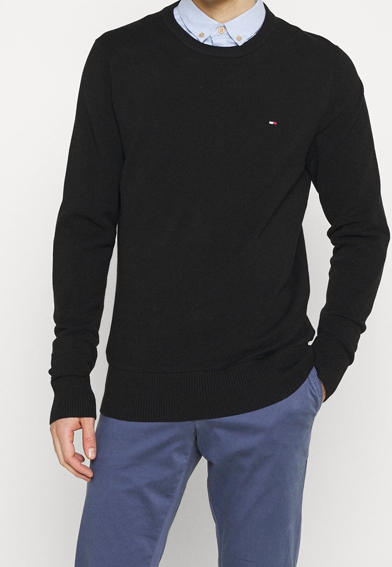 Mann in schwarzem Rundhalsausschnittpullover über einem hellblauen, knöpfbaren Hemd mit blauen Hosen, der mit einer Hand in der Tasche steht.