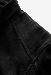 Chaqueta de ante negra con cuello alto, detalles en costuras diagonales y líneas texturizadas sutiles a lo largo del hombro para añadir interés al diseño.