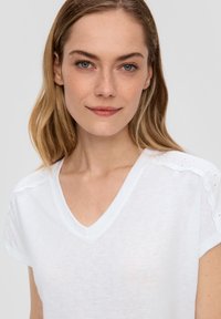 Witte v-hals t-shirt met korte mouwen, voorzien van oogjesdetails. Gemaakt van lichte stof met een licht doorschijnende textuur.