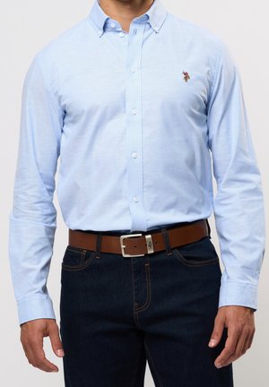 Homme portant une chemise bleu clair à boutons rentrée dans un jean bleu foncé avec une ceinture en cuir marron et un logo visible sur la poitrine de la chemise.