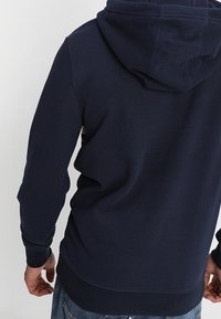 Sudadera con capucha de color azul marino, con un bolsillo tipo canguro, puños y dobladillo acanalados. Fabricada con un tejido suave y de textura lisa, adecuada para el uso casual.