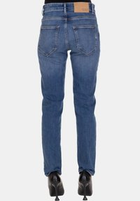 Jeans blu slim-fit con vita media, tasche posteriori e un'etichetta beige sul retro. Il tessuto ha una texture denim liscia.