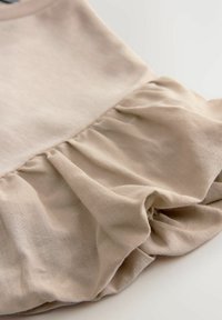 Beige bomulls-top med volangkant. Tyget ser mjukt och något texturerat ut, med en slät yta och en avslappnad design.