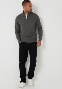 Hombre vistiendo un suéter gris oscuro con cremallera a medio abrir, pantalones negros y zapatillas blancas, de pie contra una pared blanca, mirando hacia un lado.
