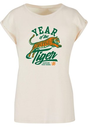 Krém színű póló rövid, felgöngyölt ujjal. Zöld és narancssárga grafikával ellátott szöveg: "TIGRIS ÉVE" és egy élénk tigris illusztráció.