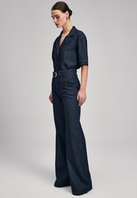 Tuta in denim scuro con maniche corte, chiusura frontale con bottoni e pantaloni a gamba larga; presenta accessori in oro e dettagli di cucitura a contrasto.