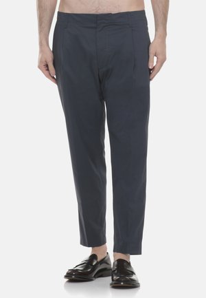 Uomo che indossa pantaloni sartoriali blu scuro e mocassini in pelle nera, in piedi contro uno sfondo bianco uniforme.