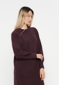 Burgundy kötött pulóver kerek nyakkivágással és hosszú ujjal, sima textúrával és laza fazonnal, a comb közepéig ér.