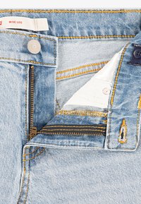Nærbilde av lyseblå vide jeans med glidelås, knapp og merke. Fremhever søm detaljer og stofftekstur.
