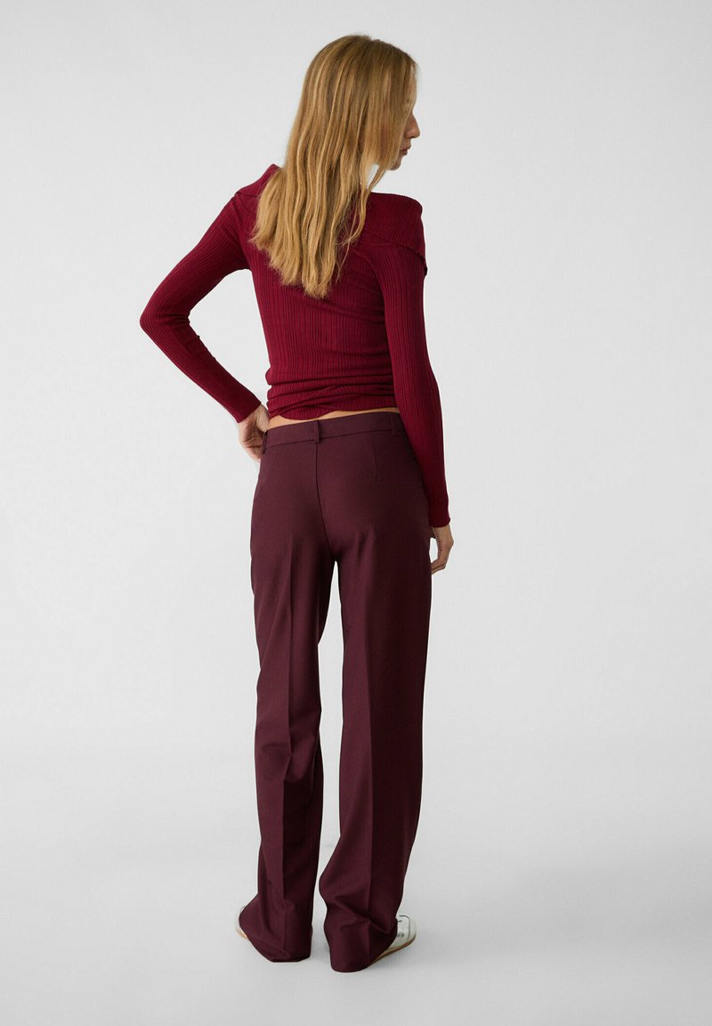 Donna con lunghi capelli biondi, che indossa un top a spalle scoperte a coste color bordeaux e pantaloni abbinati color bordeaux, in piedi davanti a uno sfondo grigio uniforme.