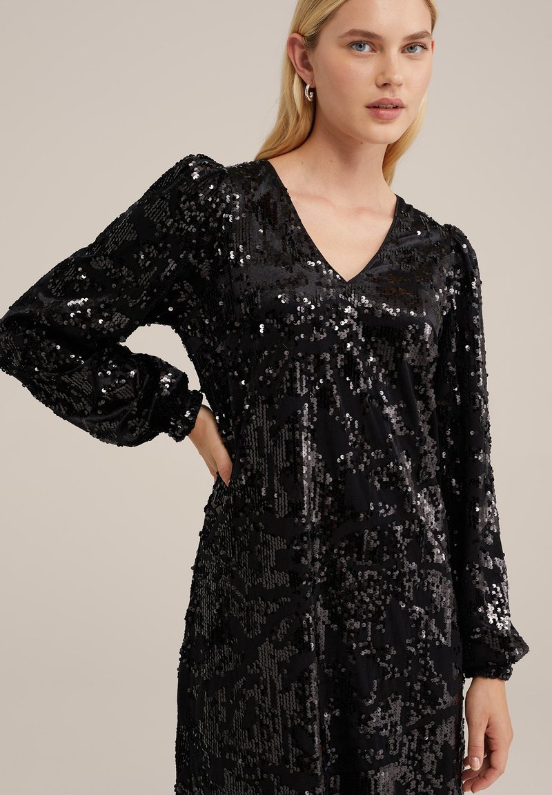 Schwarzes Paillettenkleid mit V-Ausschnitt, langen Puffärmeln und strukturiertem Muster. Der Stoff ist glänzend und hat aufgrund der Pailletten eine reflektierende Qualität.