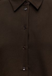 Chemise en tissu marron foncé avec un col pointu et des boutons noirs sur le devant au centre.