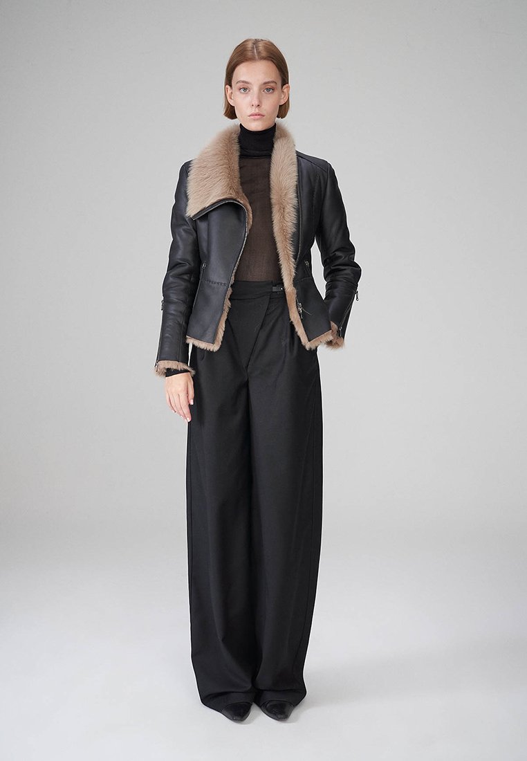 Schwarze Lederjacke mit einem beigen Kunstfellkragen, kombiniert mit hoch geschnittenen schwarzen Hosen. Glatte Textur und figurbetontes Design.