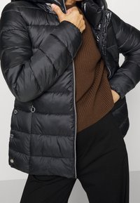 Černá péřová bunda s kapucí. Má lesklý povrch, prošívaný design, stříbrné kovové doplňky a přední zip. Nositelná přes hnědý svetr.