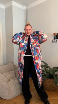 Pikowany płaszcz w kwiatowy wzór z jasnoczerwonymi, niebieskimi i zielonymi motywami, z prążkowanymi mankietami, przednim zamkiem błyskawicznym i luźnym krojem. Pod spodem czarne spodnie.