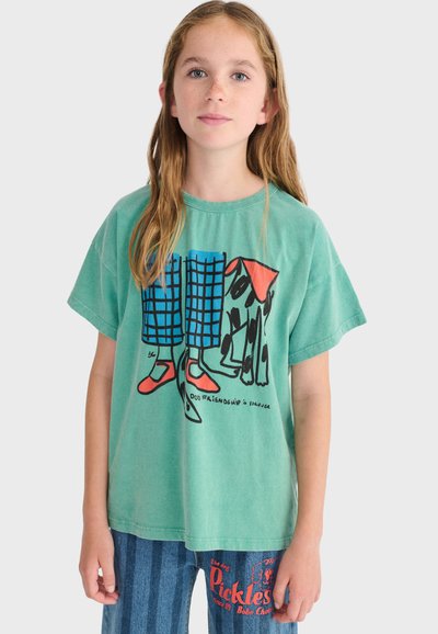 Mlada djevojka s dugom kosom nosi zelenu majicu s crtanim nogama i psom, uparenu s plavim hlačama na pruge s crvenim natpisom.