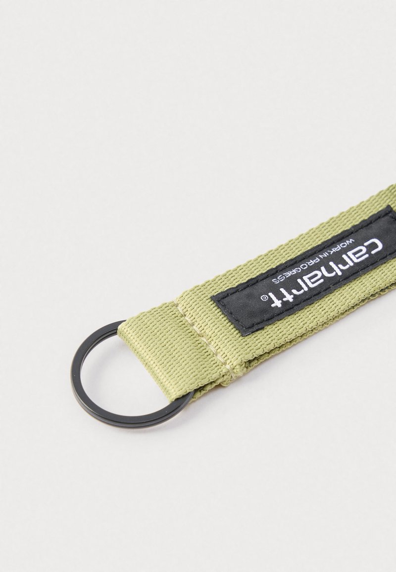 Portachiavi in tessuto verde intrecciato con anello metallico nero e etichetta nera con la scritta "Carhartt Work In Progress" su sfondo bianco.